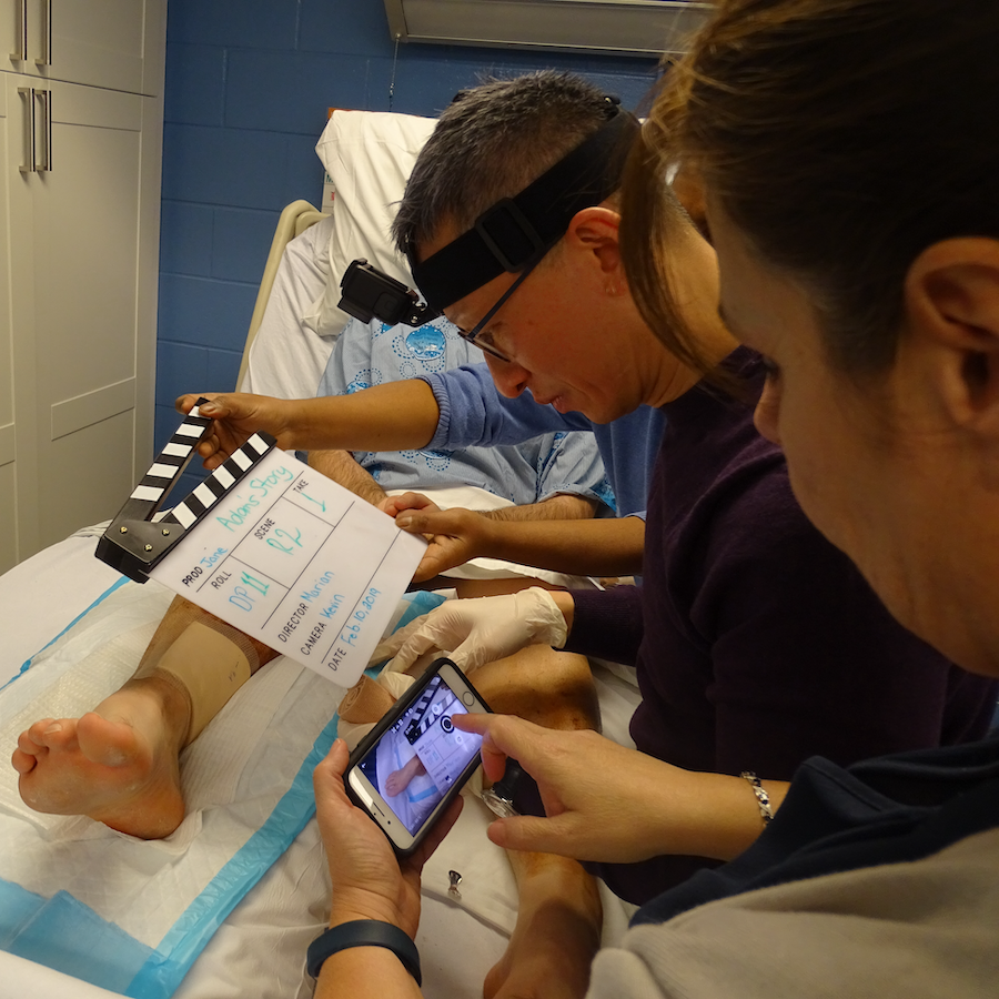 Dr. Kevin Woo leaning over a simulation model manikin, ready to begin working, while another person holds up a cellphone camera to film on his left and someone on his right holds a film clapper over the manikin